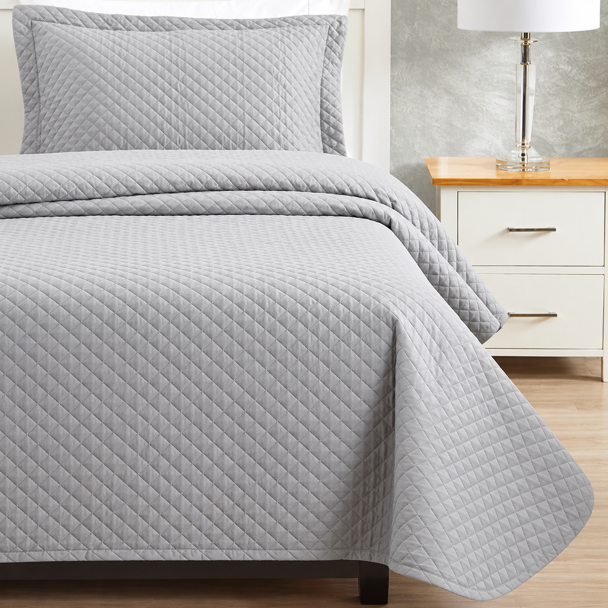 Pictured: Bedroom setting with grey bedding on a white bedframe, next to a matching nightstand with a table lamp. The bedding features the Radiance Diamond Quilt Bedspread, which has a diamond stitch pattern that adds texture to the blanket.