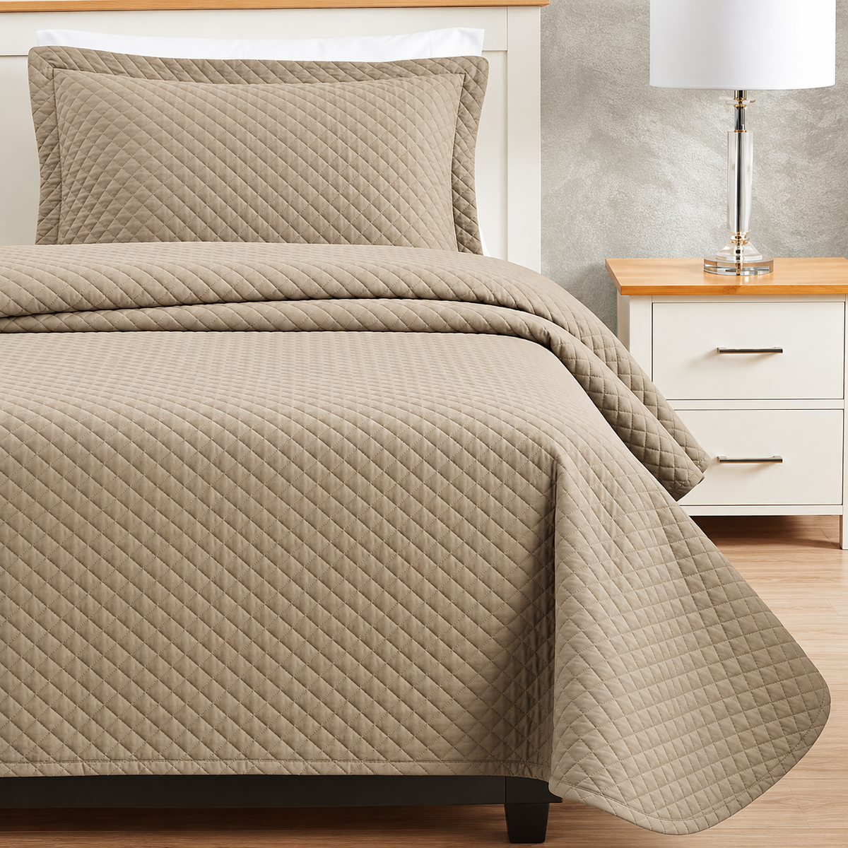 Pictured: Bedroom setting with tan bedding on a white bedframe, next to a matching nightstand with a table lamp. The bedding features the Radiance Diamond Quilt Bedspread, which has a diamond stitch pattern that adds texture to the blanket.