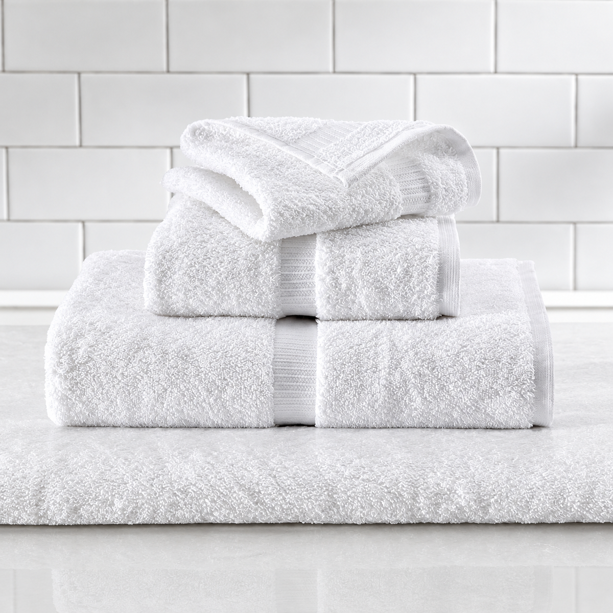 Pictured: Different white towels neatly folded and stacked on top of each other. They sit on a marble surface in front of white subway tile. The towels feature a vertical dobby border.