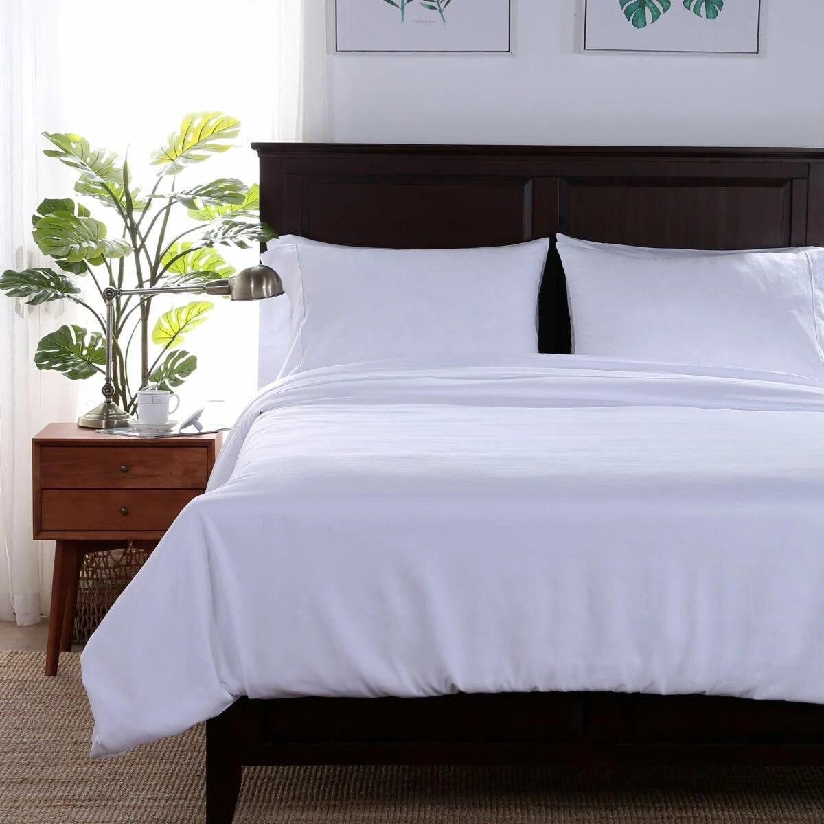 Pictured: Modern bedroom setting with a nightstand holding a plant, teacup, and silver lamp on the left. On the right is a dark wood bedframe with white bedding and pillows.