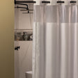 Pictured: Hookless Shower Curtain hanging from a black rod in a standing shower. The curtain is white with a clear window at the top. There are 2 thin black stripes that create a border around the curtain.