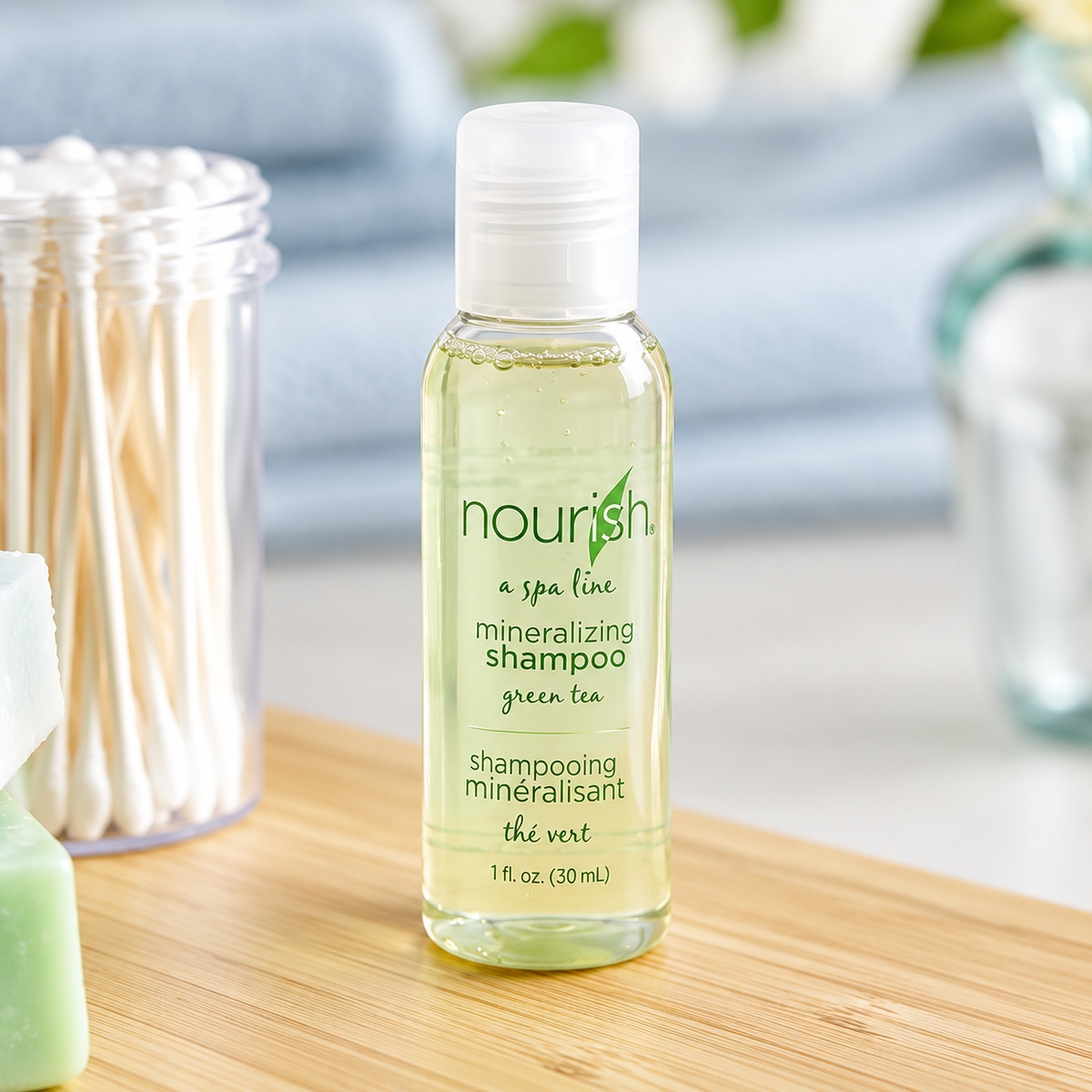 Pictured: Small standing clear bottle  holding gel-like liquid with a clear cap in the forefront of a bathroom setting. The bottle is part of a spa line called Nourish, this product is the mineralizing shampoo with green tea. The bottle holds 1 fluid oz. (30 mL)