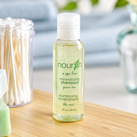 Pictured: Small standing clear bottle  holding gel-like liquid with a clear cap in the forefront of a bathroom setting. The bottle is part of a spa line called Nourish, this product is the mineralizing shampoo with green tea. The bottle holds 1 fluid oz. (30 mL)