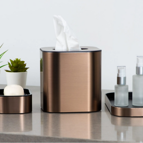 Bronze tissue box and other bathroom accessories on a counter with a white background