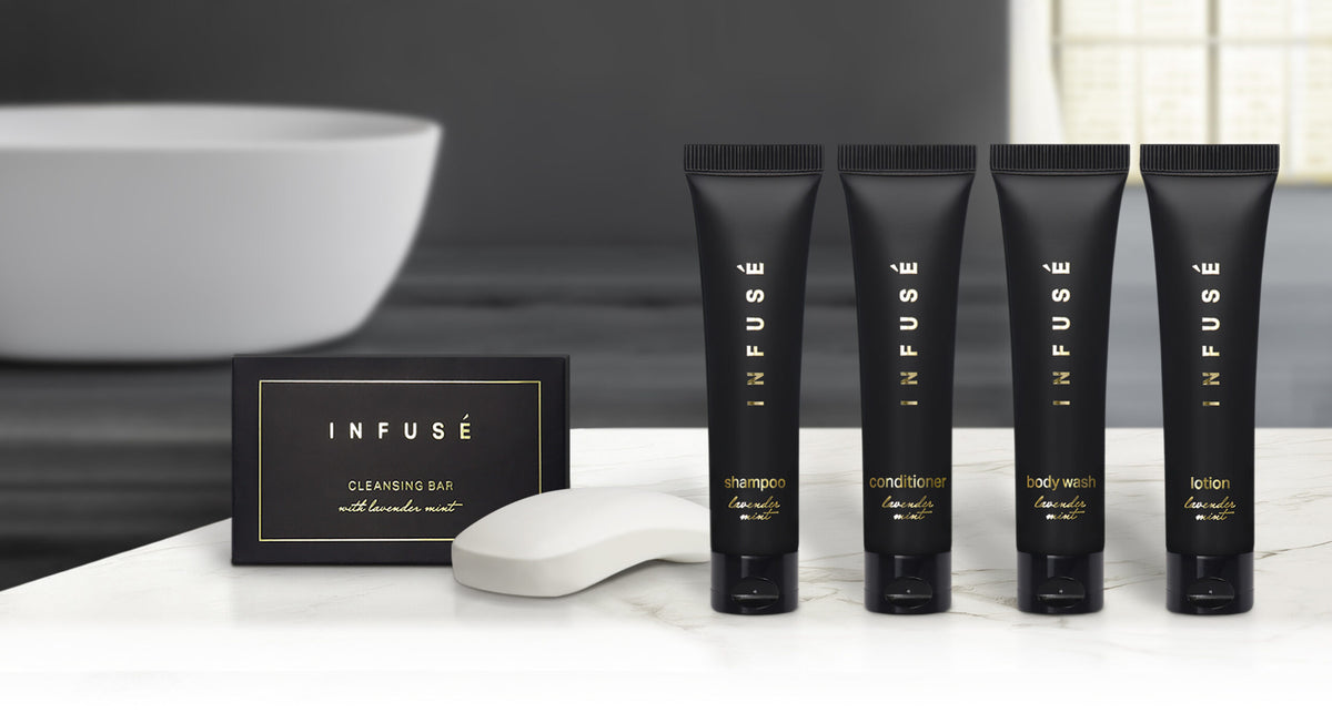 Pictured: Infuse Amenity collection lined up on a white marble counter with a bathtub in the background. Products pictured from left to right are: Infuse Cleansing Bar, Infuse Shampoo, Infuse Conditioner, Infuse Body Wash and Infuse Lotion. All products made with lavender mint. The cleansing bar comes in a black cardboard box with gold accents. Every other product pictured comes in a black squeeze tube with gold accents and a flip cap.