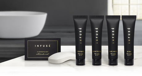 Pictured: Infuse Amenity collection lined up on a white marble counter with a bathtub in the background. Products pictured from left to right are: Infuse Cleansing Bar, Infuse Shampoo, Infuse Conditioner, Infuse Body Wash and Infuse Lotion. All products made with lavender mint. The cleansing bar comes in a black cardboard box with gold accents. Every other product pictured comes in a black squeeze tube with gold accents and a flip cap.