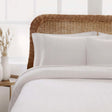 White textured bedding set with two white pillows and a matching white quilted comforter, styled on a bed with a natural wicker headboard in a bright bedroom setting.