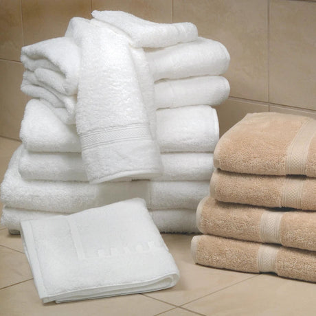 Pictured: Neatly folded stack of the Magnificence Towel collection against bathroom tile. The collection comes in the shades White (left) and Linen (right).
