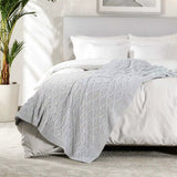 Gray textured blanket draped over a bed in a bedroom setting with a plant and framed picture.