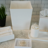 White bathroom accessories including a square vase, tissue box, and other items on a marble countertop.