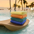 A stack of folded, vibrant colored pool towels placed on a woven mat beside a pool, with palm trees and a sunset in the background.