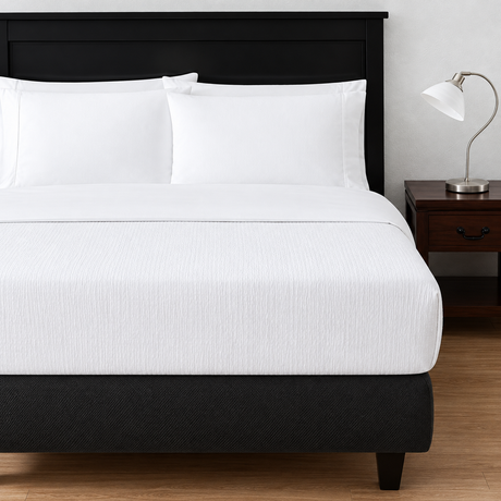 Pictured: White bedding on a dark bed frame with a nightstand holding a table lamp to the right. The bedding features the Rattan Pucker Jacquard Top Sheet which has basket-weave inspired design.