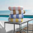 Pictured: 4 neatly folded striped beach towels sitting on a marble side table in front of a pool and the ocean. The colors from top to bottom are: Peach, Brown, Blue and Yellow.