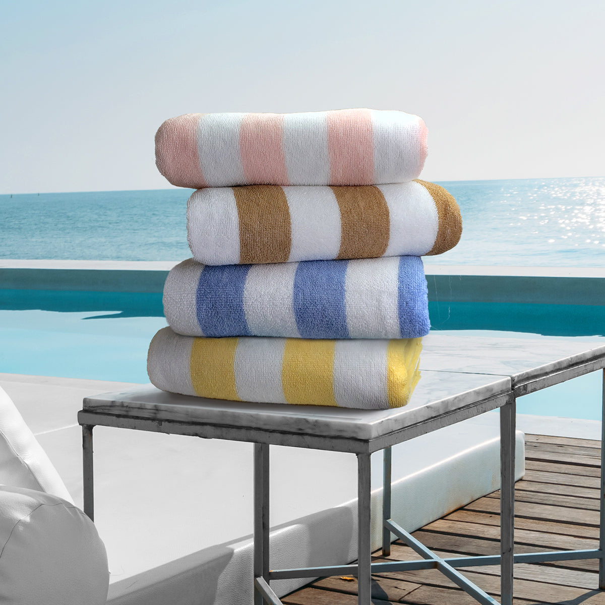 Pictured: 4 neatly folded striped beach towels sitting on a marble side table in front of a pool and the ocean. The colors from top to bottom are: Peach, Brown, Blue and Yellow.