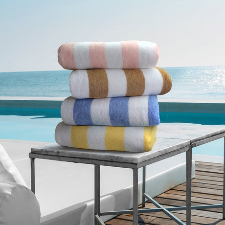 Pictured: 4 neatly folded striped beach towels sitting on a marble side table in front of a pool and the ocean. The colors from top to bottom are: Peach, Brown, Blue and Yellow.