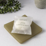 Cleansing soap bar with a white label on a light surface, accompanied by greenery.