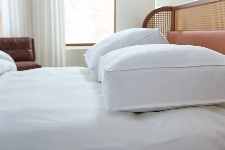 Pictured: 2 Beyond Down Side Sleeper pillows on top of a white bed with a wooden bed frame. In the background you can see a brown leather seat in front of long white drapes hanging above a window.