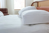 Pictured: 2 Beyond Down Side Sleeper pillows on top of a white bed with a wooden bed frame. In the background you can see a brown leather seat in front of long white drapes hanging above a window.