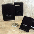 Pictured: 3 Black Makeup Washcloths against a tiled background. On the left two of the towels are draped over a  small standing towel rack, and on the right the towel is leaned against an object. The towels are embroidered to say "Makeup" in white writing.