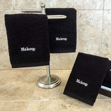 Pictured: 3 Black Makeup Washcloths against a tiled background. On the left two of the towels are draped over a  small standing towel rack, and on the right the towel is leaned against an object. The towels are embroidered to say "Makeup" in white writing.
