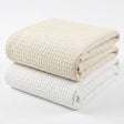 Pictured: Two waffle woven blankets neatly folded and stacked against a white background. The blanket on the bottom is in the shade "White" and the one on top is in the shade "Beige".