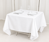 Pictured: White Cotton Craft Dynasty Tablecloth draped over a square table. The table is set with two plates, two wine glasses, two forks and two knives.