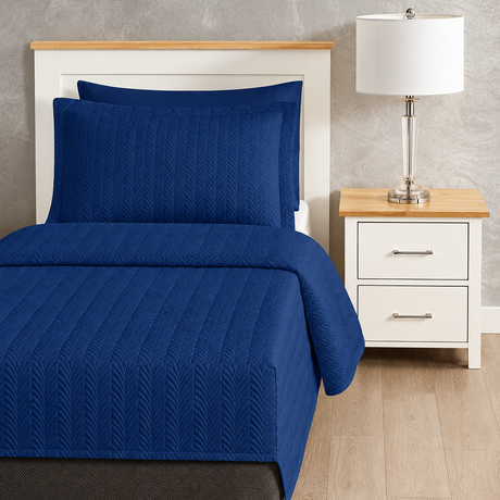 Pictured: Blue bedding on a white wooden bedframe with a matching nightstand to the right holding a glass table lamp with a white shade. The bedding is the Flourish Quilt Coverlet in the shade Sail.