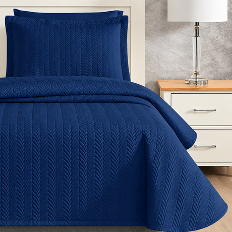 Pictured: Blue quilted bedding on a white wooden bed frame next to a matching nightstand that is holding a glass lamp with a white shade. The bedding is the Flourish Quilt Bedspread in the shade "Sail Blue".