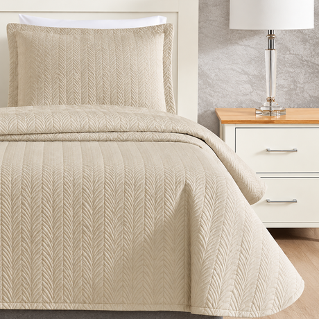 Pictured: Beige quilted bedding on a white wooden bed frame next to a matching nightstand that is holding a glass lamp with a white shade. The bedding is the Flourish Quilt Bedspread in the shade "Sand".