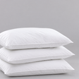 Pictured: 3 fluffy white pillows laid down and stacked on top of each other against a gray background. The product featured is the Gold Label Hospitality Pillow.