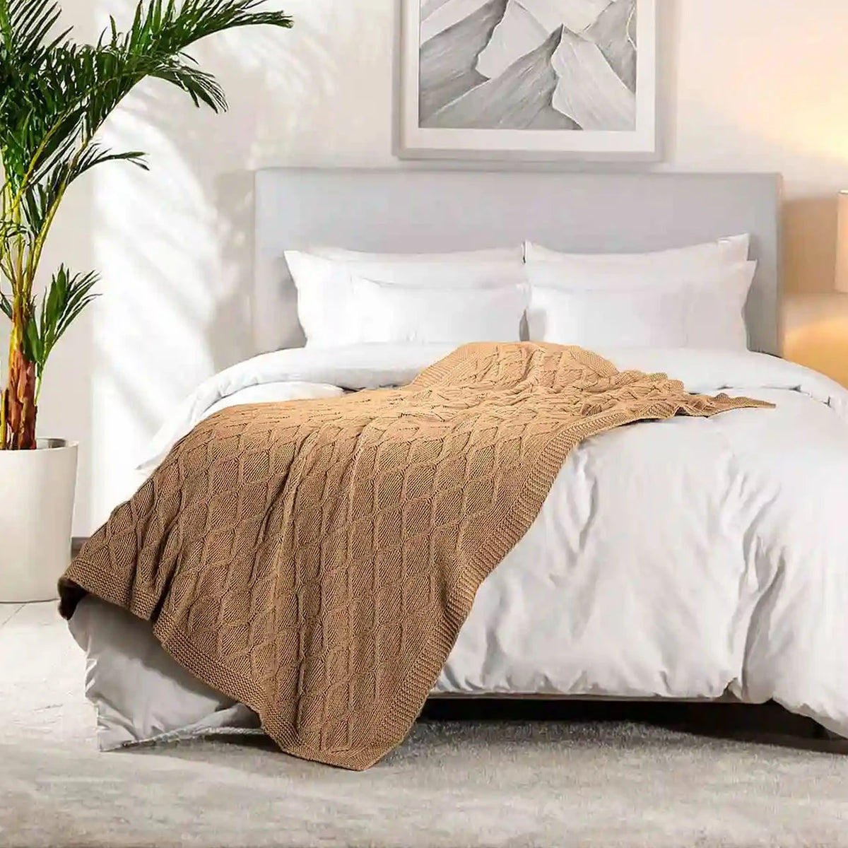 Pictured: Brown textured blanket draped over a white bed in a bedroom setting with a plant on the left and framed picture above the headboard.