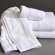 Pictured: White towels with a decorative Dobby border against a gray background. Two towels are neatly folded and stacked on the right, with another towel draped over the stack. At the top is a smaller hand towel neatly rolled.