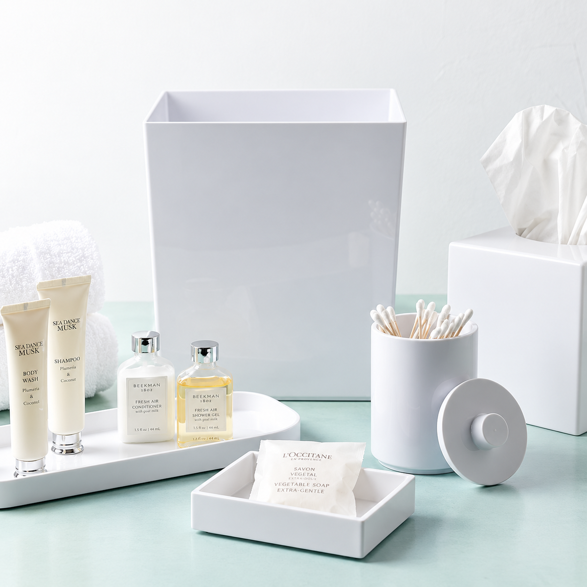 Pictured: The entire Oasis Collection on top of a sleek aqua counter. Featuring the Spa Amenity Tray holding bath and body products, the Apothecary Jar holding cotton swabs,  the Spa Boutique Tissue Box Cover, Spa Rectangular Soap Dish holding a packaged soap and the Spa Rectangular Wastebasket.