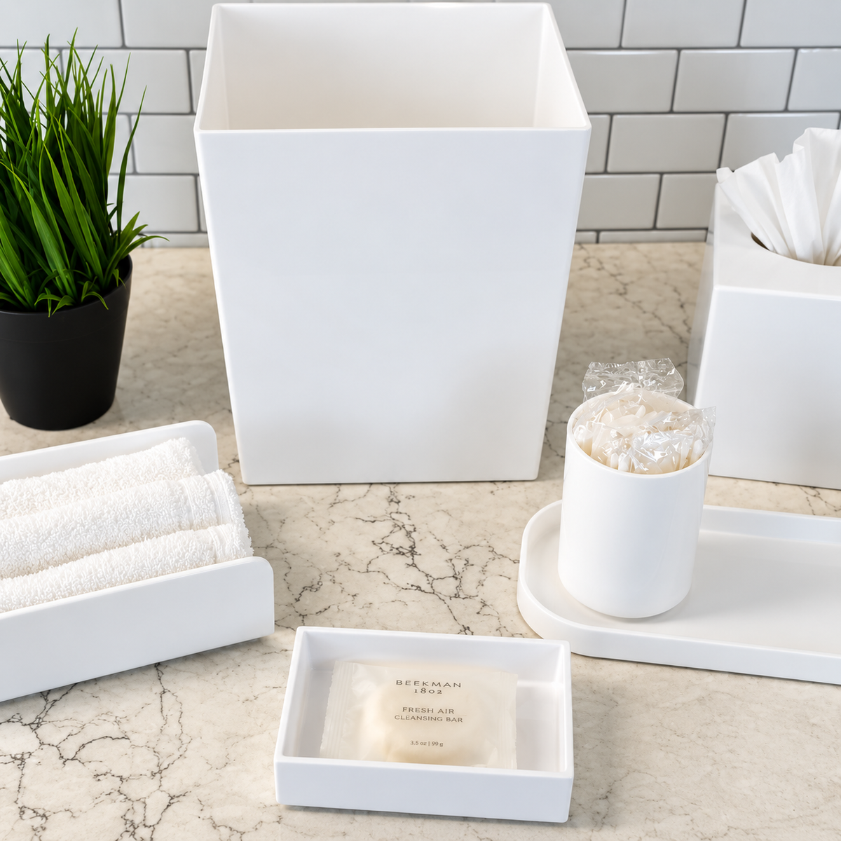 Pictured: The entire Oasis Collection in a bathroom setting. Featuring the Apothecary Jar sitting on the Spa Amenity Tray, the Spa Boutique Tissue Box Cover, Spa Rectangular Soap Dish holding a packaged Soap, the Spa Rectangular Wastebasket and Spa Towel Holder with three white towels.