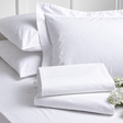 Pictured: White sheets neatly folded in front of a white pillow in a decorative pillow sham, and two white pillows sitting in the back. There are also white flowers pictured in the bottom right corner.