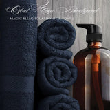 Pictured:  White cursive text that says "Oxford Magic Bleachguard" and then a serif font below that says "MAGIC BLEACHGUARD HOTEL TOWEL"
You can see 4 navy blue towels, 3 of which are neatly rolled and stacked while the other one is standing on the left. There is a brown dispenser bottle to the right.
