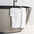 Pictured: Modern white bathroom with honeycomb tile and a stainless steel tub. Draped over the tub are 3 white towels from the Oxford Signature Towel Collection.