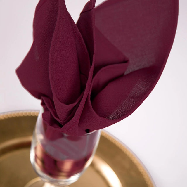 Pictured: Decorative napkin in the shade New Burgundy folded fancifully inside of a wine glass on a golden plate.