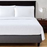 Pictured: Bedroom setting with a dark wood frame and matching nightstand. The white bed features the Jacquard top sheet, which has a naturally scrunched or wrinkled texture in little box stitches.