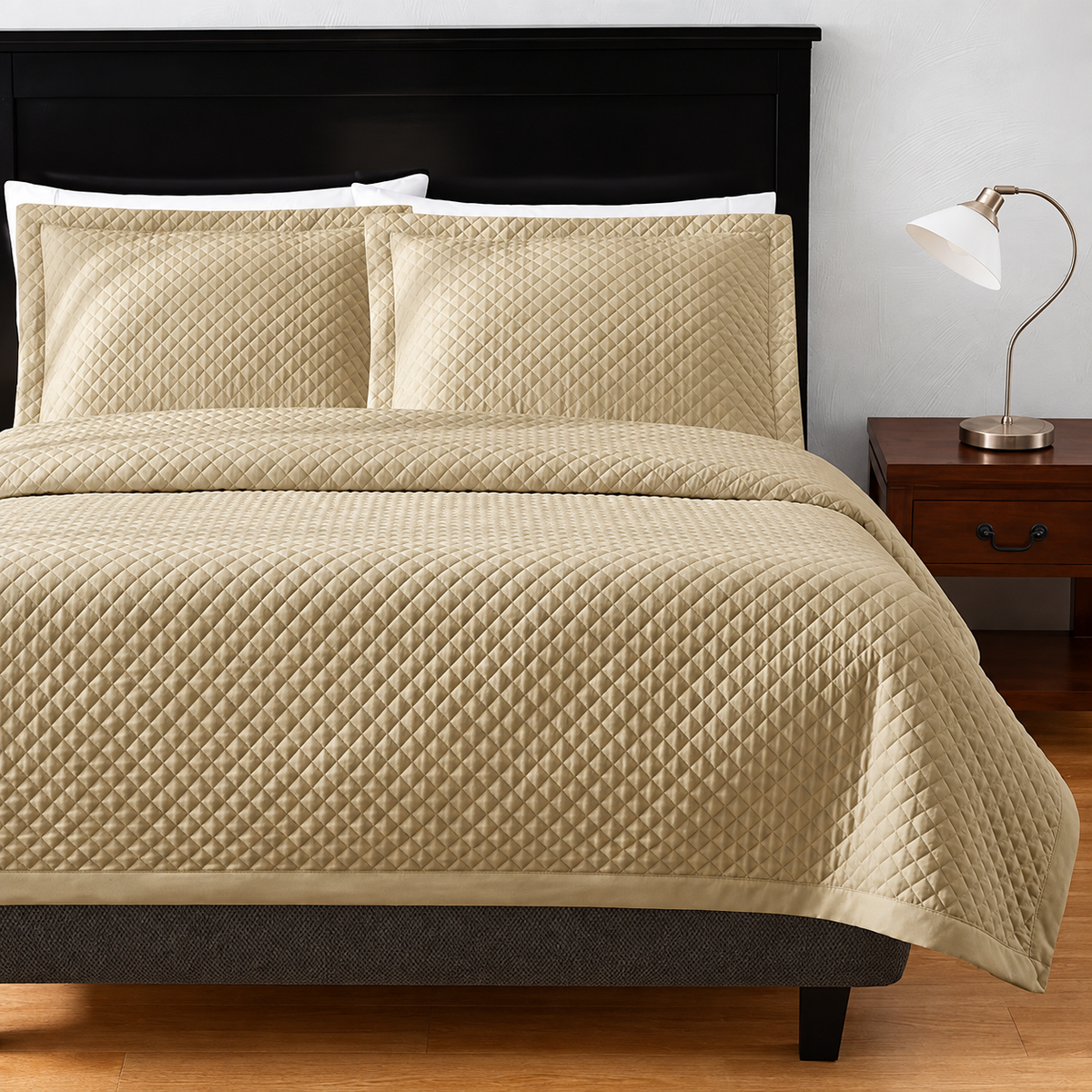 Pictured: Bedroom setting with beige bedding on a dark wood frame. The bedding features the Radiance Diamond Coverlet, which has a diamond stitch pattern and adds a texture to the blanket. To the right of the bed is a dark wood nightstand with a silver lamp on top.