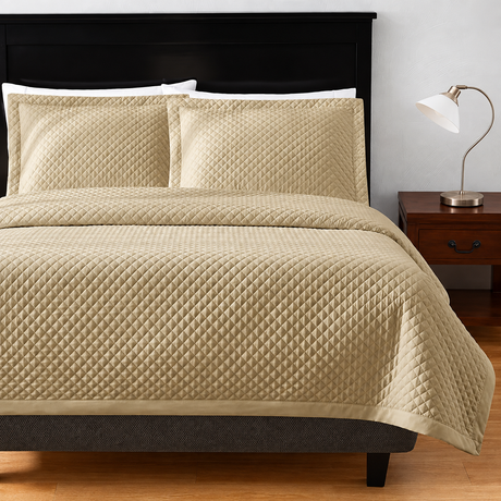 Pictured: Bedroom setting with beige bedding on a dark wood frame. The bedding features the Radiance Diamond Coverlet, which has a diamond stitch pattern and adds a texture to the blanket. To the right of the bed is a dark wood nightstand with a silver lamp on top.