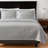 Pictured: Bedroom setting with grey bedding on a dark wood frame. The bedding features the Radiance Diamond Coverlet, which has a diamond stitch pattern and adds a texture to the blanket. To the right of the bed is a dark wood nightstand with a silver lamp on top.