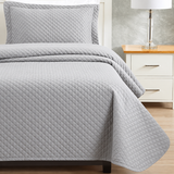 Pictured: Bedroom setting with grey bedding on a white bedframe, next to a matching nightstand with a table lamp. The bedding features the Radiance Diamond Quilt Bedspread, which has a diamond stitch pattern that adds texture to the blanket.