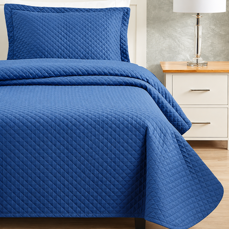 Pictured: Bedroom setting with blue bedding on a white bedframe, next to a matching nightstand with a table lamp. The bedding features the Radiance Diamond Quilt Bedspread, which has a diamond stitch pattern that adds texture to the blanket.