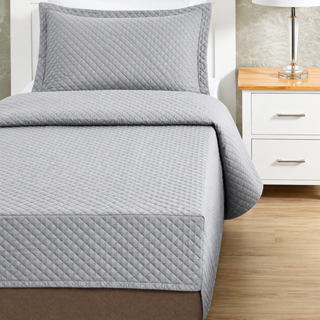 Pictured: Grey colored bedding on a white bedframe next to a matching nightstand (right) which is holding a lamp. The bedding features the Radiance Quilt Fitted Coverlet. It has a diamond stitch, creating a textured pattern.