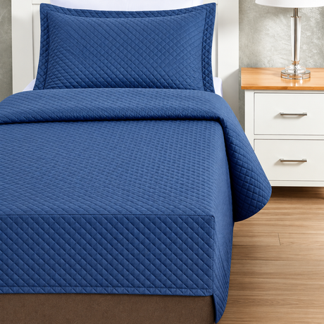 Pictured: Blue colored bedding on a white bedframe next to a matching nightstand (right) which is holding a lamp. The bedding features the Radiance Quilt Fitted Coverlet. It has a diamond stitch, creating a textured pattern.