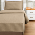 Pictured: Tan colored bedding on a white bedframe next to a matching nightstand (right) which is holding a lamp. The bedding features the Radiance Quilt Fitted Coverlet. It has a diamond stitch, creating a textured pattern.