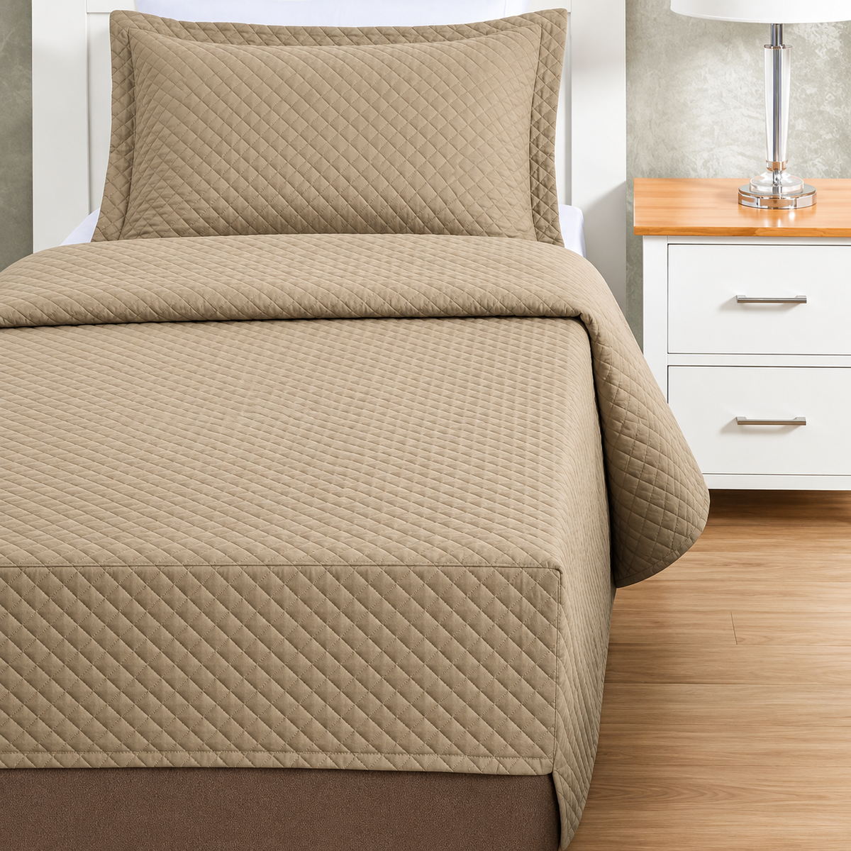 Pictured: Tan colored bedding on a white bedframe next to a matching nightstand (right) which is holding a lamp. The bedding features the Radiance Quilt Fitted Coverlet. It has a diamond stitch, creating a textured pattern.