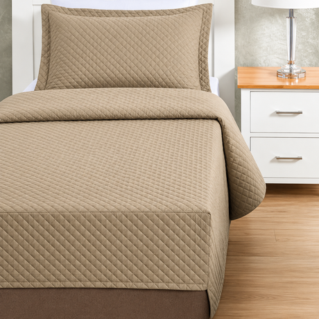Pictured: Tan colored bedding on a white bedframe next to a matching nightstand (right) which is holding a lamp. The bedding features the Radiance Quilt Fitted Coverlet. It has a diamond stitch, creating a textured pattern.