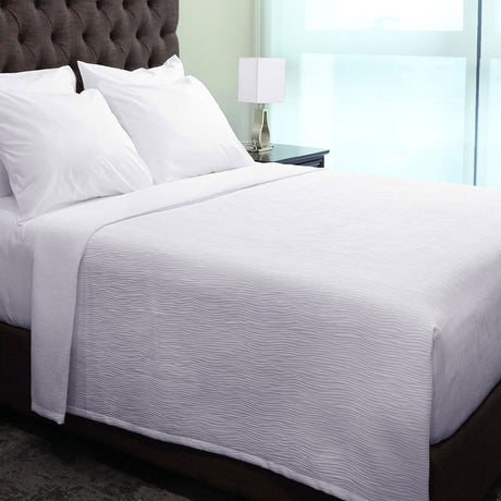Pictured: White bedding pictured atop a dark fabric bed frame. In the background is a nightstand, a table lamp, and some sheer curtains covering large windows. The bed features the Ripple Weave Puff Jacquard Top Sheet, which has a wavy texture/pattern.
